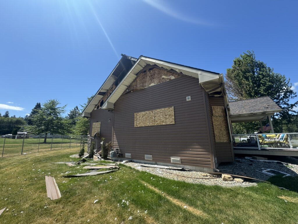 Damaged home with boarded windows and missing siding, set against a blue sky. Debris scattered on the lawn; nearby fencing is visible.