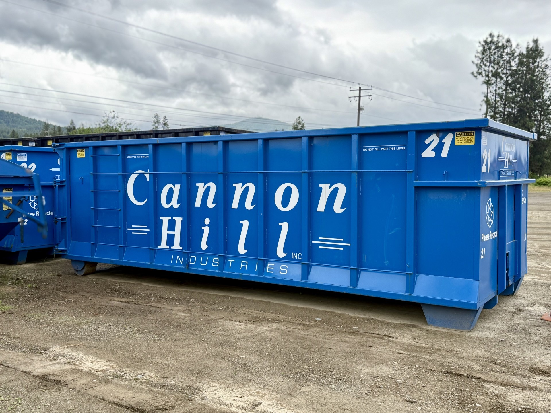 Blue dumpster labeled "Cannon Hill Industries" with a caution sign, set against a cloudy sky and surrounded by gravel. Designed for waste disposal.