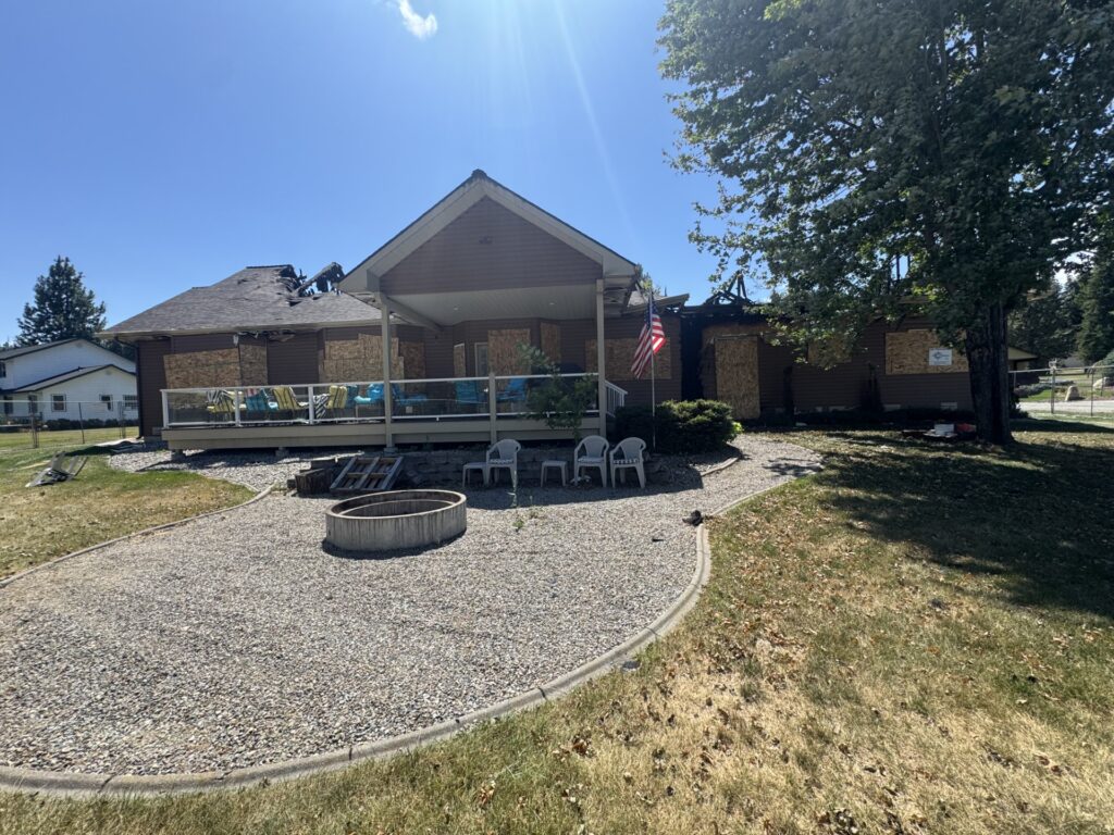 Home with significant fire damage, showing boarded windows and a damaged roof. The front yard features a gravel area and a fire pit.