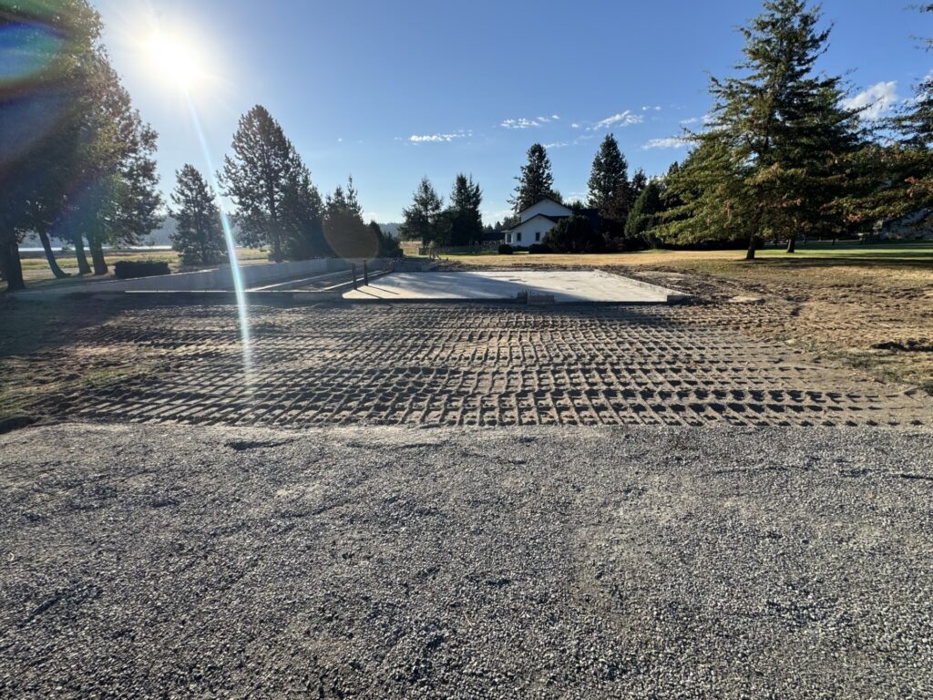 Construction site showing a newly poured concrete slab with tire tracks on a partially cleared area, framed by trees and a house in the background.